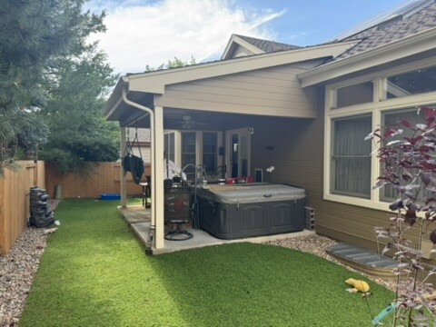 A backyard with artificial grass, a covered patio, and a hot tub. Surrounded by a wooden fence with trees, the setting is peaceful under a partly cloudy sky.