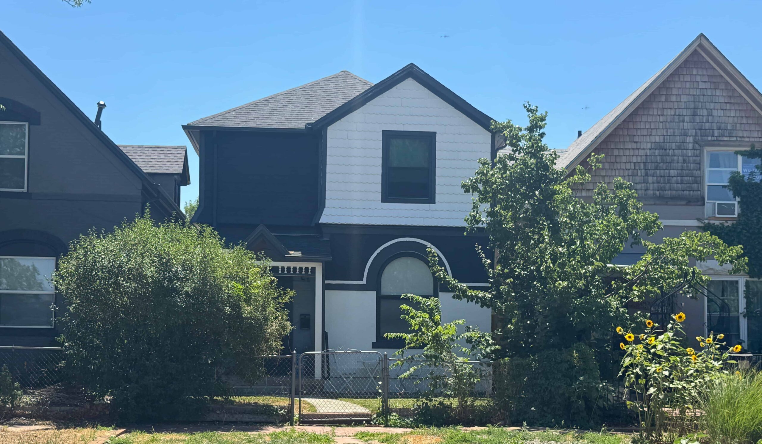 A two-story house with a mix of dark and light siding, framed by trees and sunflowers, under a clear blue sky, conveying a serene suburban feel.