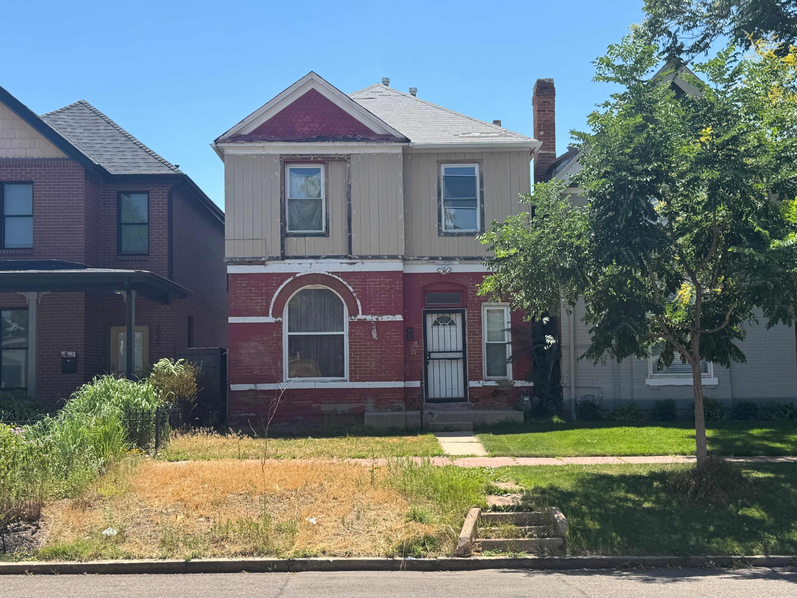 A two-story brick house with a weathered facade, arched window, and door. The yard is partly overgrown and sunlit. A tree provides partial shade.