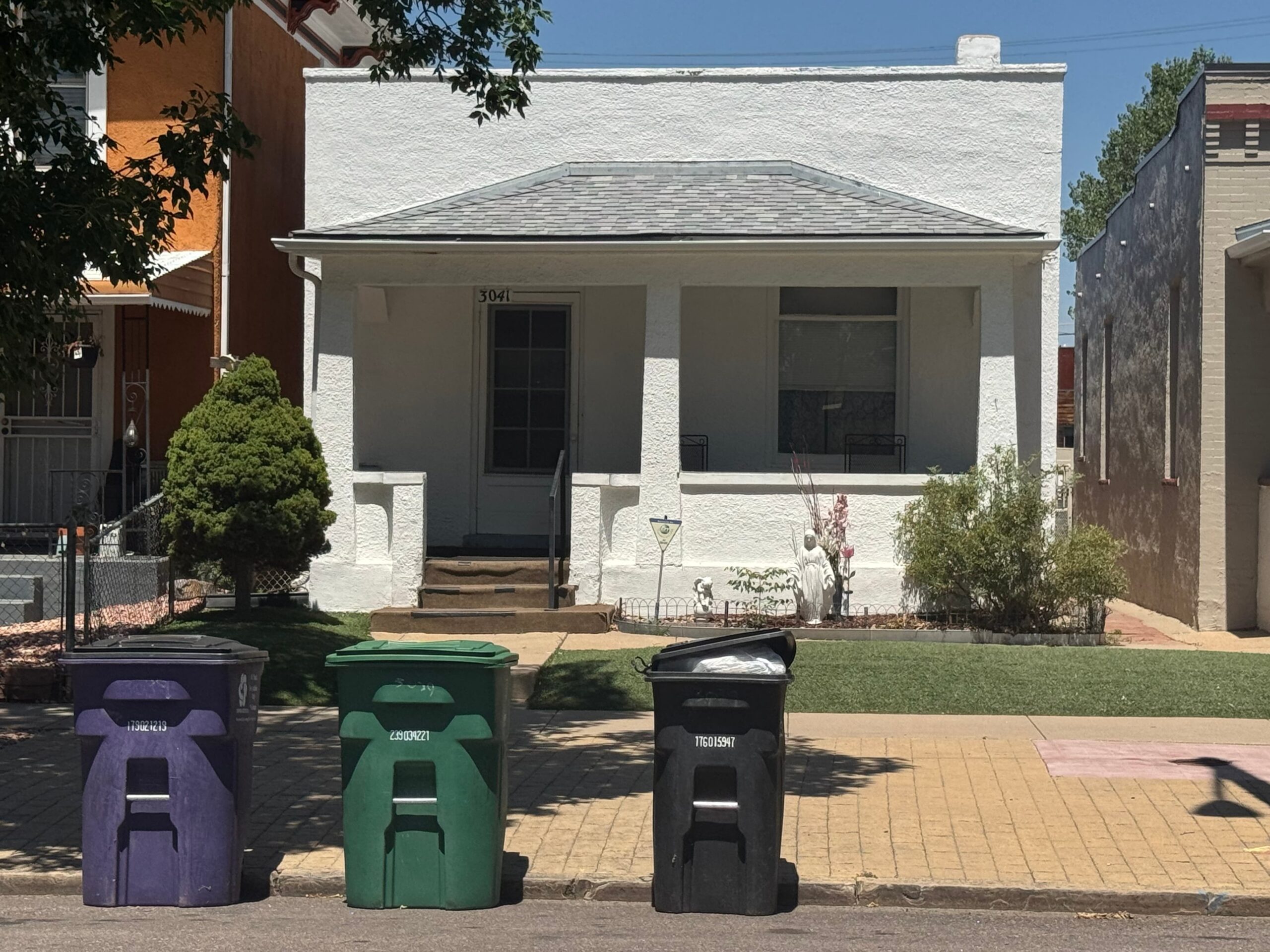 A small white house with a gray roof and two front windows sits behind a neat lawn. Three trash bins—purple, green, and black—are lined up in front.
