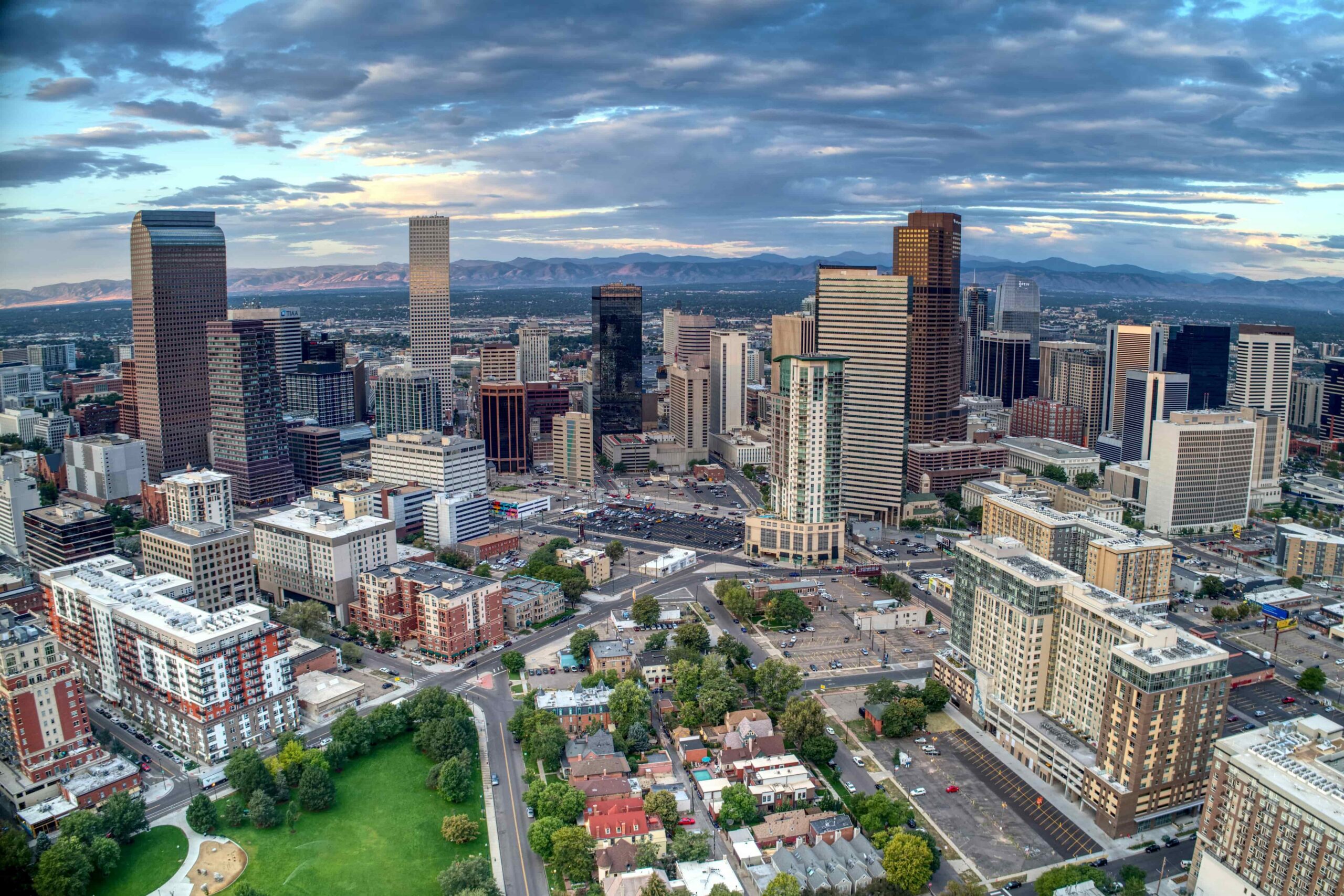 Aerial view of a bustling city skyline with tall buildings under a cloudy sky. Green parks and residential areas are visible in the foreground. Vibrant and expansive.