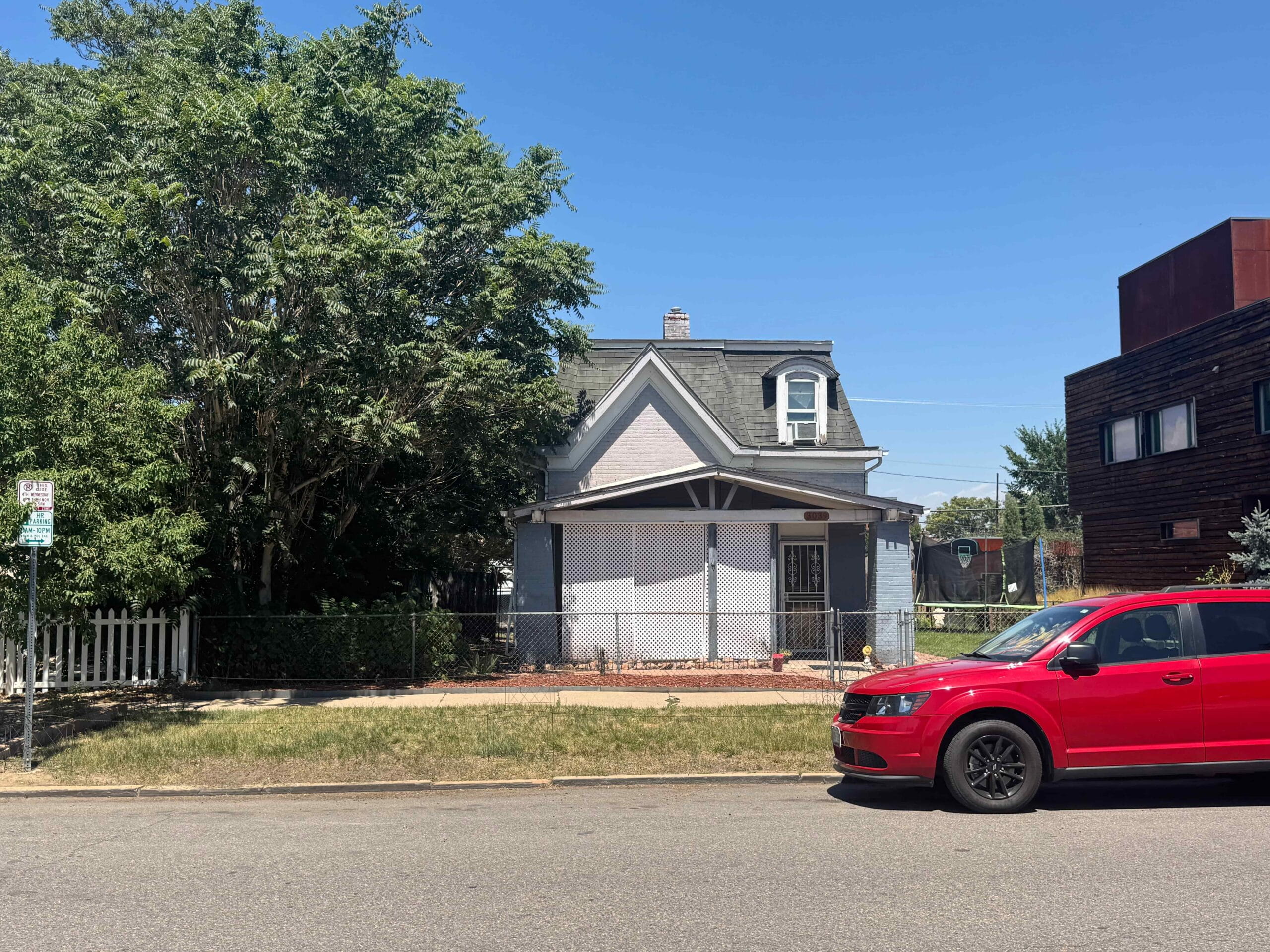 A small house with a gabled roof stands between a large tree and a modern building. A red car is parked on the street in front. Bright, sunny day.