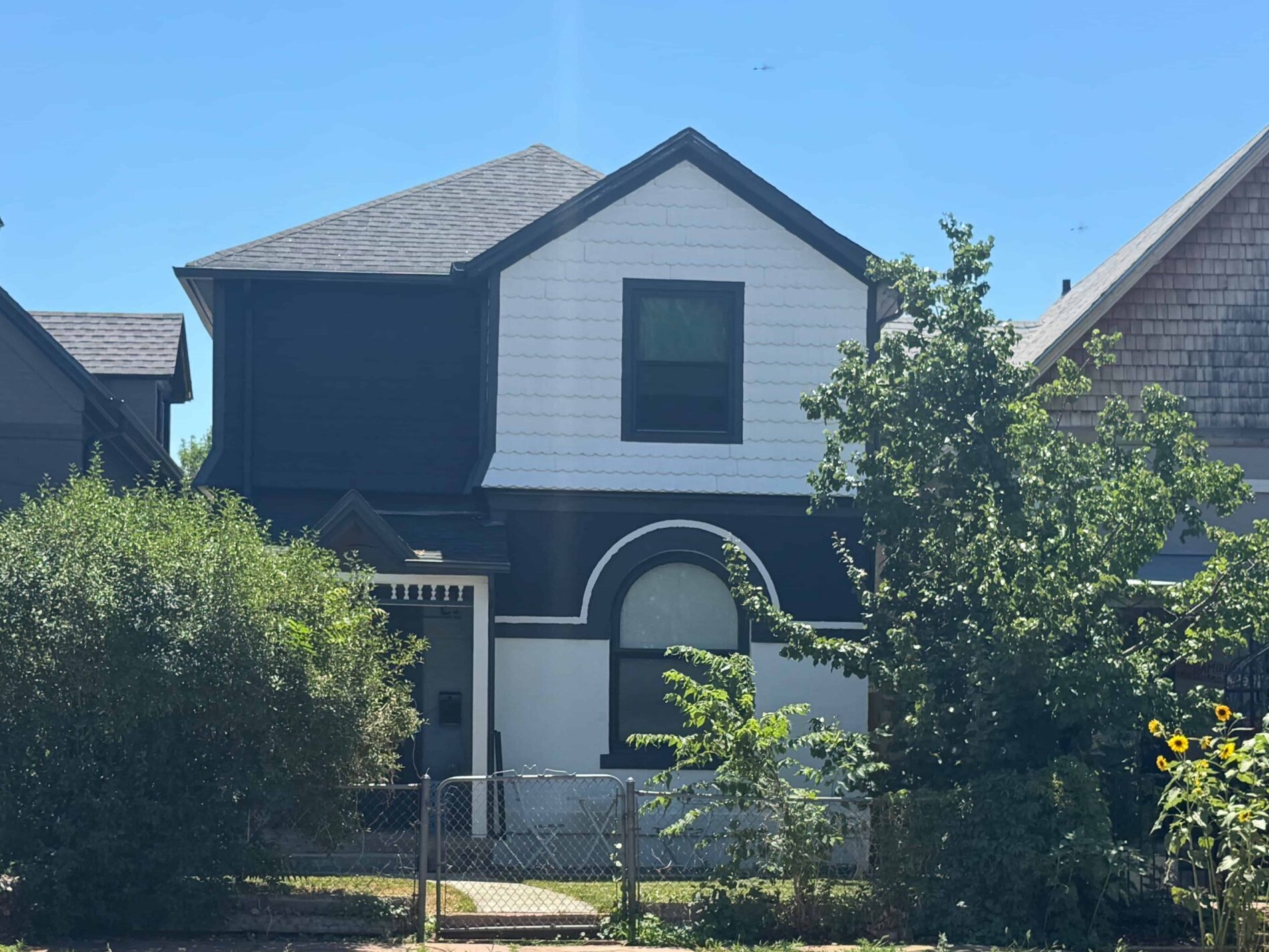 A two-story house with a mix of dark and light siding, framed by trees and sunflowers, under a clear blue sky, conveying a serene suburban feel.