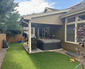 A backyard with artificial grass, a covered patio, and a hot tub. Surrounded by a wooden fence with trees, the setting is peaceful under a partly cloudy sky.
