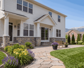 A two-story house with stone and white siding exterior. A stone path leads to a porch, surrounded by lush flowers and a manicured lawn under a clear sky.