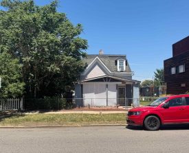A small house with a gabled roof stands between a large tree and a modern building. A red car is parked on the street in front. Bright, sunny day.