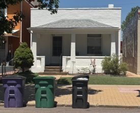 A small white house with a gray roof and two front windows sits behind a neat lawn. Three trash bins—purple, green, and black—are lined up in front.
